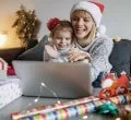 Mãe e filha sorridentes, usando gorro de Natal, sentadas na mesa, olhando juntas o que está na tela de um notebook.