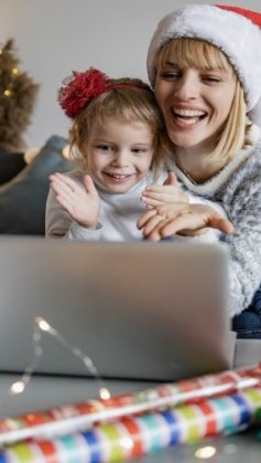 Mãe e filha sorridentes, usando gorro de Natal, sentadas na mesa, olhando juntas o que está na tela de um notebook.
