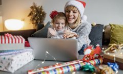 Mãe e filha sorridentes, usando gorro de Natal, sentadas na mesa, olhando juntas o que está na tela de um notebook.