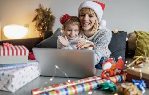 Mãe e filha sorridentes, usando gorro de Natal, sentadas na mesa, olhando juntas o que está na tela de um notebook.