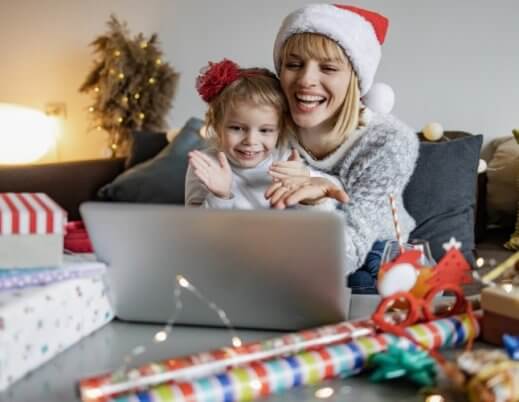 Mãe e filha sorridentes, usando gorro de Natal, sentadas na mesa, olhando juntas o que está na tela de um notebook.
