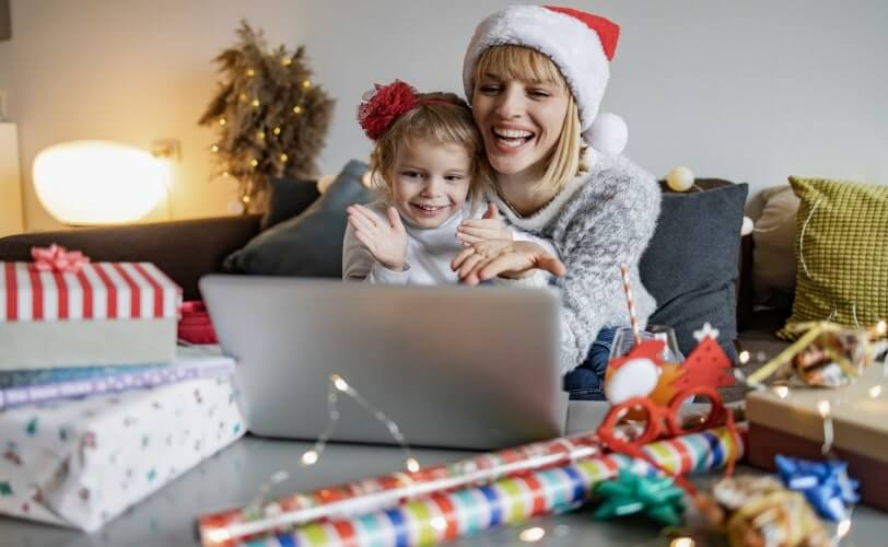 Mãe e filha sorridentes, usando gorro de Natal, sentadas na mesa, olhando juntas o que está na tela de um notebook.