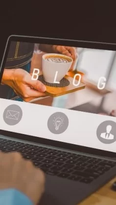 Pessoa usando um notebook em uma mesa de madeira, com a tela exibindo a palavra “BLOG” e a foto de uma xícara de café sendo servida. A pessoa segura um copo de café na mão esquerda enquanto digita com a direita. Um smartphone está apoiado ao lado do notebook.