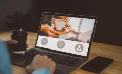 Pessoa usando um notebook em uma mesa de madeira, com a tela exibindo a palavra “BLOG” e a foto de uma xícara de café sendo servida. A pessoa segura um copo de café na mão esquerda enquanto digita com a direita. Um smartphone está apoiado ao lado do notebook.