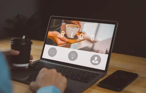 Pessoa usando um notebook em uma mesa de madeira, com a tela exibindo a palavra “BLOG” e a foto de uma xícara de café sendo servida. A pessoa segura um copo de café na mão esquerda enquanto digita com a direita. Um smartphone está apoiado ao lado do notebook.