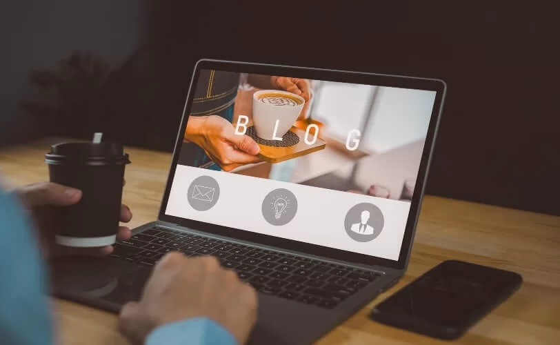 Pessoa usando um notebook em uma mesa de madeira, com a tela exibindo a palavra “BLOG” e a foto de uma xícara de café sendo servida. A pessoa segura um copo de café na mão esquerda enquanto digita com a direita. Um smartphone está apoiado ao lado do notebook.