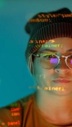 A imagem mostra uma pessoa com óculos e gorro, observando atentamente uma tela com linhas de código projetadas sobre seu rosto, criando um efeito visual que une o humano à tecnologia. A cena simboliza a imersão no mundo da programação, onde criatividade e lógica se encontram. As cores azuladas e o reflexo do monitor reforçam o ambiente digital e concentrado, representando o cotidiano de profissionais de tecnologia, desenvolvedores e cientistas de dados. Essa composição destaca a importância da inteligência humana na construção do futuro tecnológico.