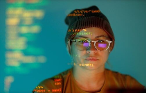A imagem mostra uma pessoa com óculos e gorro, observando atentamente uma tela com linhas de código projetadas sobre seu rosto, criando um efeito visual que une o humano à tecnologia. A cena simboliza a imersão no mundo da programação, onde criatividade e lógica se encontram. As cores azuladas e o reflexo do monitor reforçam o ambiente digital e concentrado, representando o cotidiano de profissionais de tecnologia, desenvolvedores e cientistas de dados. Essa composição destaca a importância da inteligência humana na construção do futuro tecnológico.