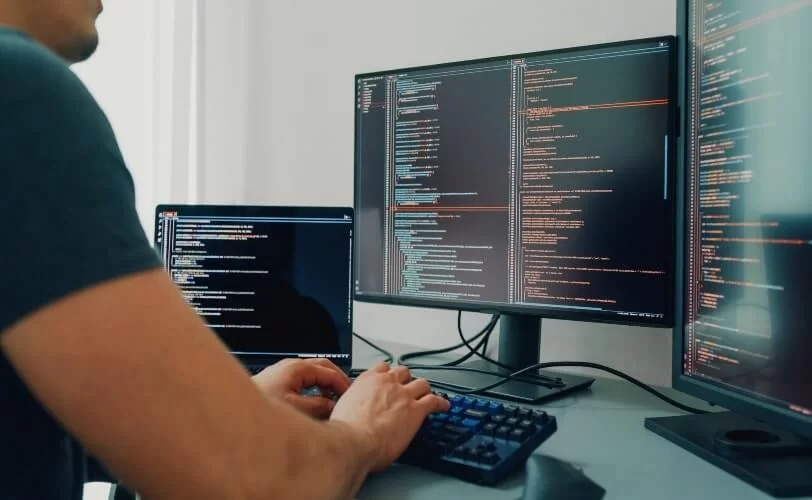 A imagem mostra uma pessoa digitando em um teclado mecânico, com vários monitores exibindo linhas de código em destaque. O ambiente transmite foco e produtividade, típico de programadores e engenheiros de software trabalhando em projetos complexos. As telas exibem código em diferentes janelas e cores, representando a organização de múltiplas tarefas e sistemas. A iluminação suave e o enquadramento lateral reforçam a atmosfera técnica e concentrada, simbolizando o trabalho analítico e criativo por trás da tecnologia, segurança digital e inovação em desenvolvimento de software.