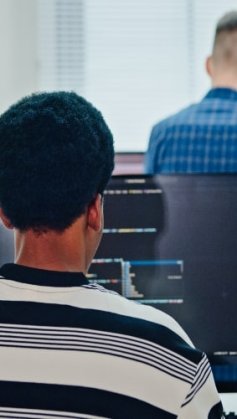 A imagem mostra uma pessoa programando em frente a múltiplos monitores, com linhas de código na tela, enquanto outro colega aparece ao fundo. O ambiente transmite foco, colaboração e inovação, típicos de equipes de desenvolvedores de software e profissionais de tecnologia. O uso de fones de ouvido e laptops reforça a ideia de concentração e imersão em um projeto digital, representando o dinamismo e a sinergia que impulsionam o desenvolvimento de soluções tecnológicas e a transformação digital nas empresas modernas.