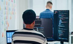 A imagem mostra uma pessoa programando em frente a múltiplos monitores, com linhas de código na tela, enquanto outro colega aparece ao fundo. O ambiente transmite foco, colaboração e inovação, típicos de equipes de desenvolvedores de software e profissionais de tecnologia. O uso de fones de ouvido e laptops reforça a ideia de concentração e imersão em um projeto digital, representando o dinamismo e a sinergia que impulsionam o desenvolvimento de soluções tecnológicas e a transformação digital nas empresas modernas.