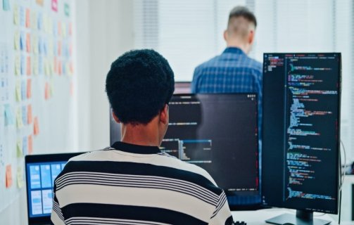 A imagem mostra uma pessoa programando em frente a múltiplos monitores, com linhas de código na tela, enquanto outro colega aparece ao fundo. O ambiente transmite foco, colaboração e inovação, típicos de equipes de desenvolvedores de software e profissionais de tecnologia. O uso de fones de ouvido e laptops reforça a ideia de concentração e imersão em um projeto digital, representando o dinamismo e a sinergia que impulsionam o desenvolvimento de soluções tecnológicas e a transformação digital nas empresas modernas.