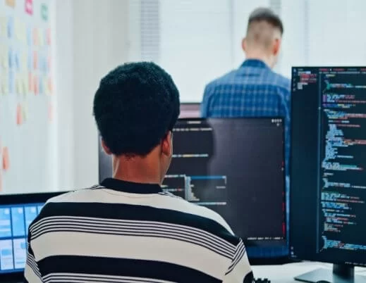 A imagem mostra uma pessoa programando em frente a múltiplos monitores, com linhas de código na tela, enquanto outro colega aparece ao fundo. O ambiente transmite foco, colaboração e inovação, típicos de equipes de desenvolvedores de software e profissionais de tecnologia. O uso de fones de ouvido e laptops reforça a ideia de concentração e imersão em um projeto digital, representando o dinamismo e a sinergia que impulsionam o desenvolvimento de soluções tecnológicas e a transformação digital nas empresas modernas.
