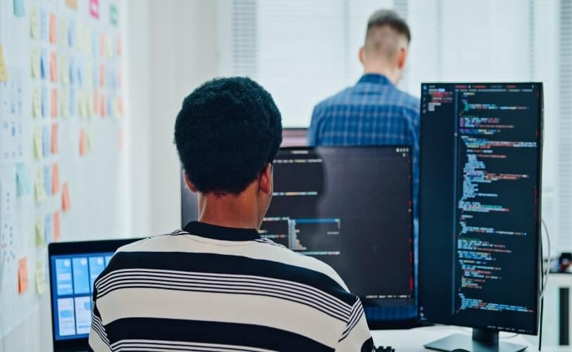 A imagem mostra uma pessoa programando em frente a múltiplos monitores, com linhas de código na tela, enquanto outro colega aparece ao fundo. O ambiente transmite foco, colaboração e inovação, típicos de equipes de desenvolvedores de software e profissionais de tecnologia. O uso de fones de ouvido e laptops reforça a ideia de concentração e imersão em um projeto digital, representando o dinamismo e a sinergia que impulsionam o desenvolvimento de soluções tecnológicas e a transformação digital nas empresas modernas.