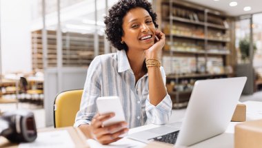 Empreendedora sorrindo enquanto mexe no celular, com laptop aberto em ambiente de loja física com estoque ao fundo
